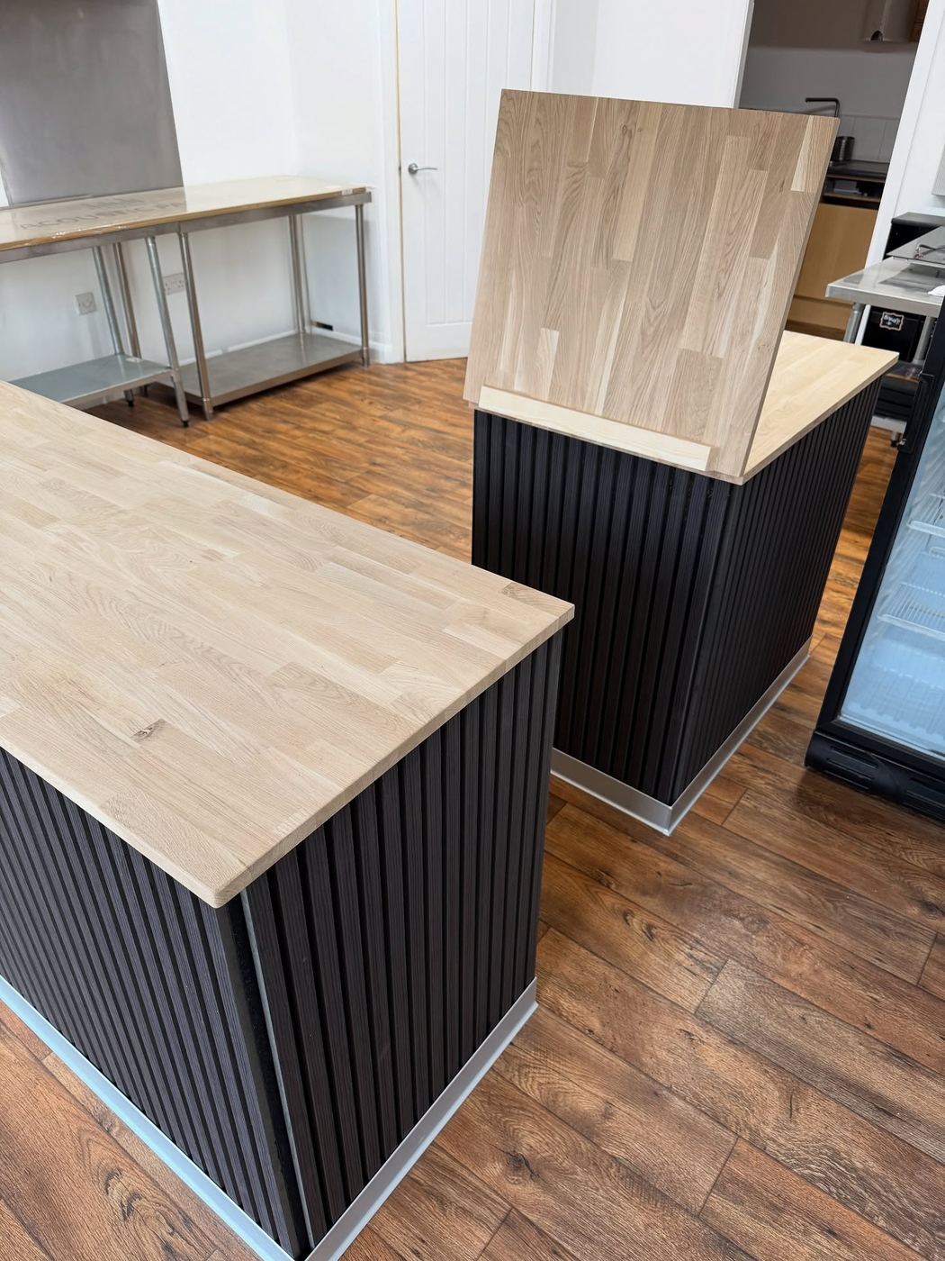 Two counters clad in dark fluted panelling with thick solid oak tops, one top propped upright